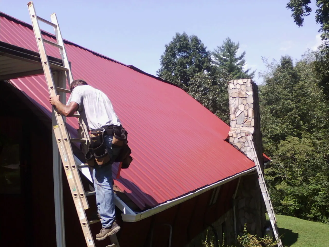 Roofer on a ladder beside a red metal roof and stone chimney — Soffit & Fascia Repair by Roofing Brothers in San Andreas